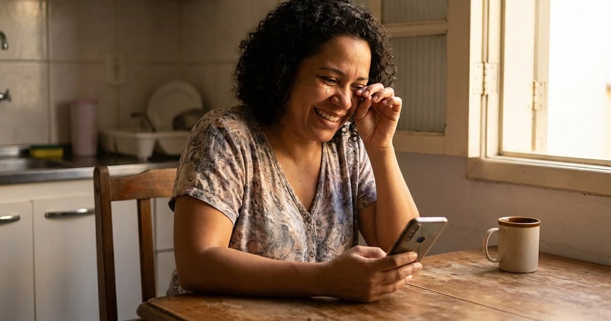Mulher brasileira adulta sorrindo ao olhar o celular na cozinha de casa, aliviada — restituição automática cashback do Imposto de Renda 2026 paga via Pix CPF
