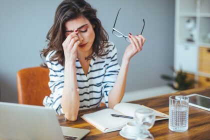 Mulher com expressão de cansaço, massageando a testa enquanto segura seus óculos, sentada em frente a um laptop em um escritório doméstico.