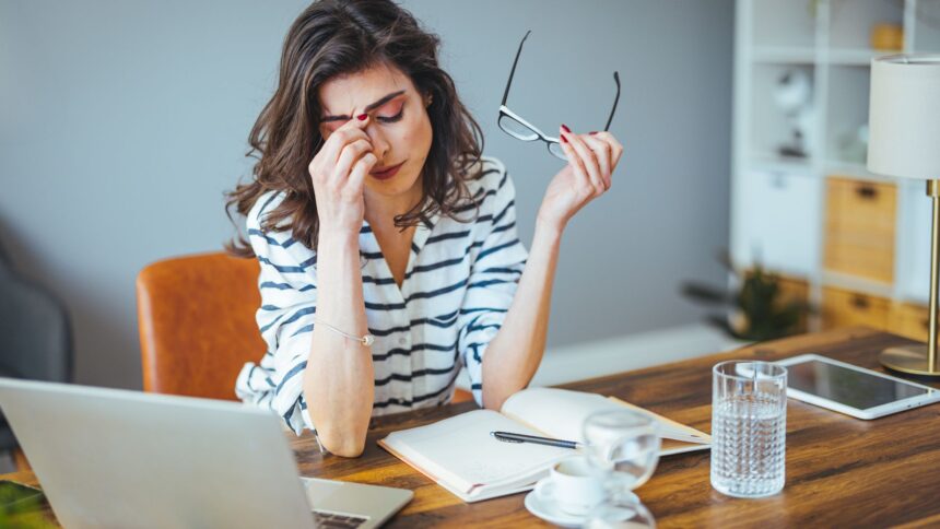 Mulher com expressão de cansaço, massageando a testa enquanto segura seus óculos, sentada em frente a um laptop em um escritório doméstico.