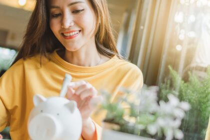 Mulher sorrindo enquanto coloca dinheiro em um cofrinho, representando o ato de economizar e gerenciar finanças pessoais.