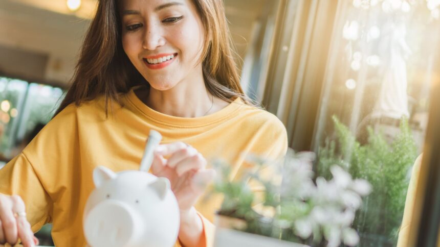 Mulher sorrindo enquanto coloca dinheiro em um cofrinho, representando o ato de economizar e gerenciar finanças pessoais.